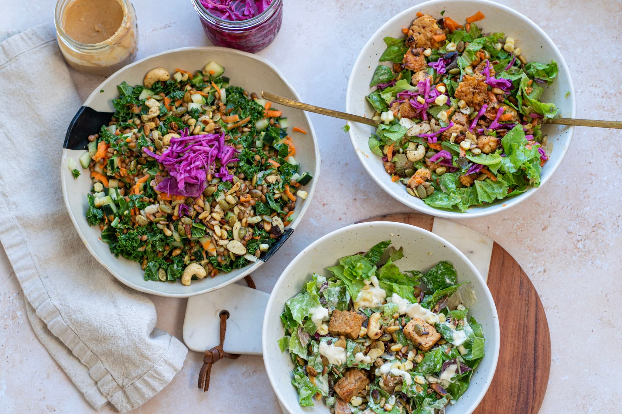 Three whole-food salad bowls on a stone counter — kale with cashews, butter lettuce with tempeh, and a creamy caesar